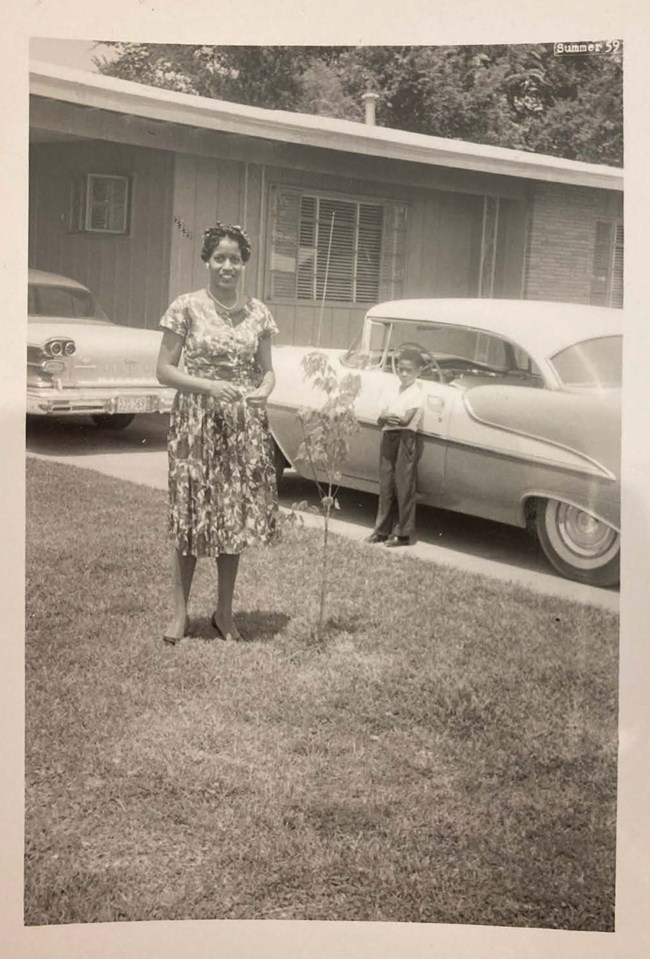 Myrlie Evers in a summer dress stands beside a young tree in short grass. Behind, young Darrell Evers leans with arms crossed against a 1950s-style car in a driveway of a ranch house. Sepia-toned.