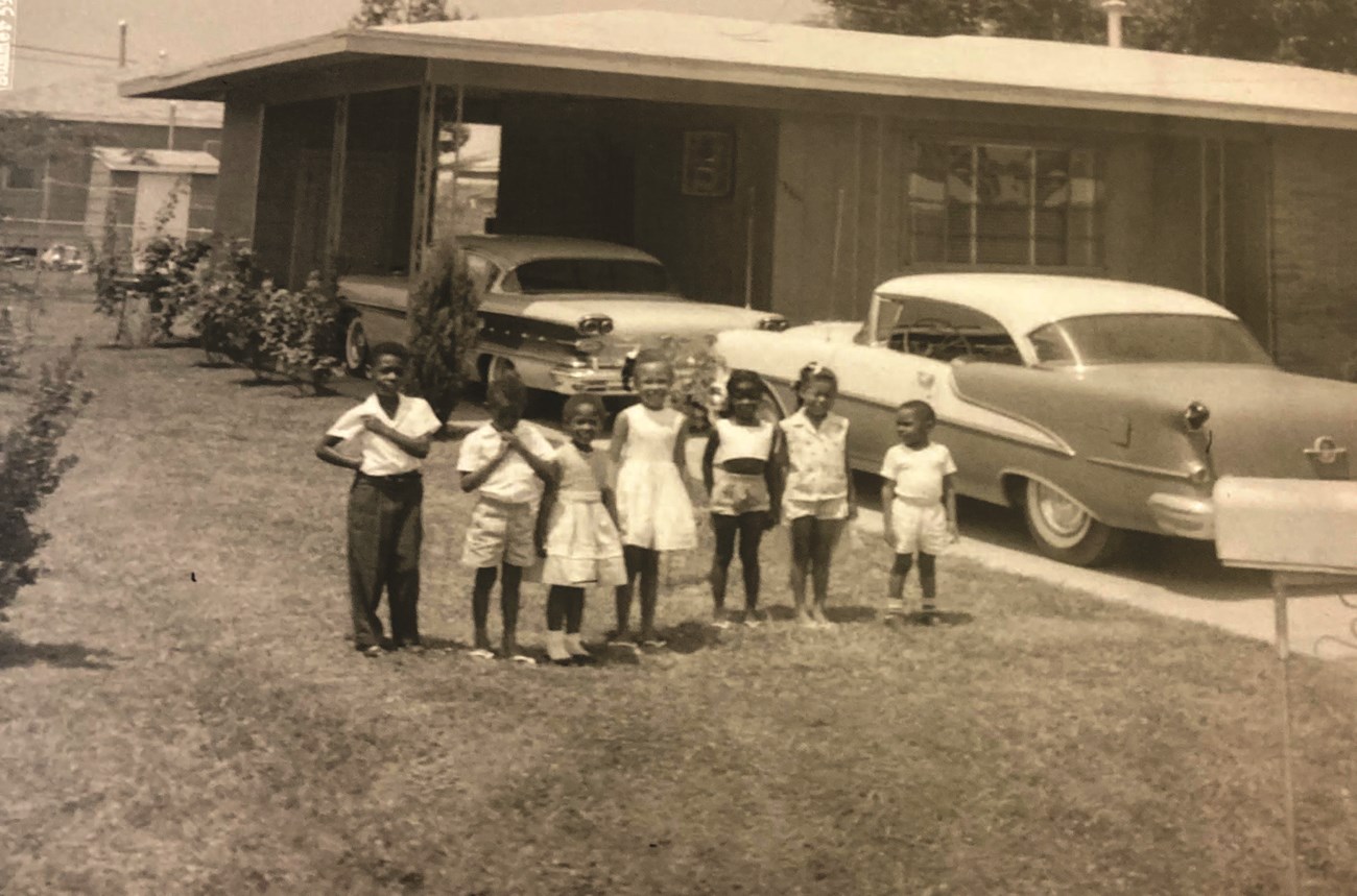 Seven African American children stand side-by-side in the grass beside two 1950s-era cars in the driveway of a ranch house, sepia-toned photo.