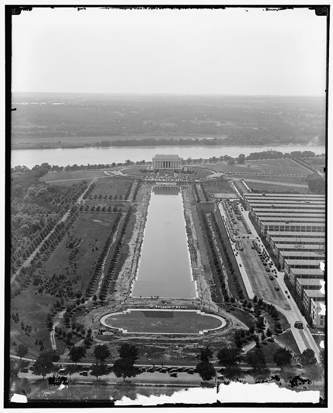 High angle view toward Lincoln Memorial in 1922 where people gather on the steps. It includes the rectangular reflecting pool, tree-lined roads and paths, and the Tidal Basin beyond.