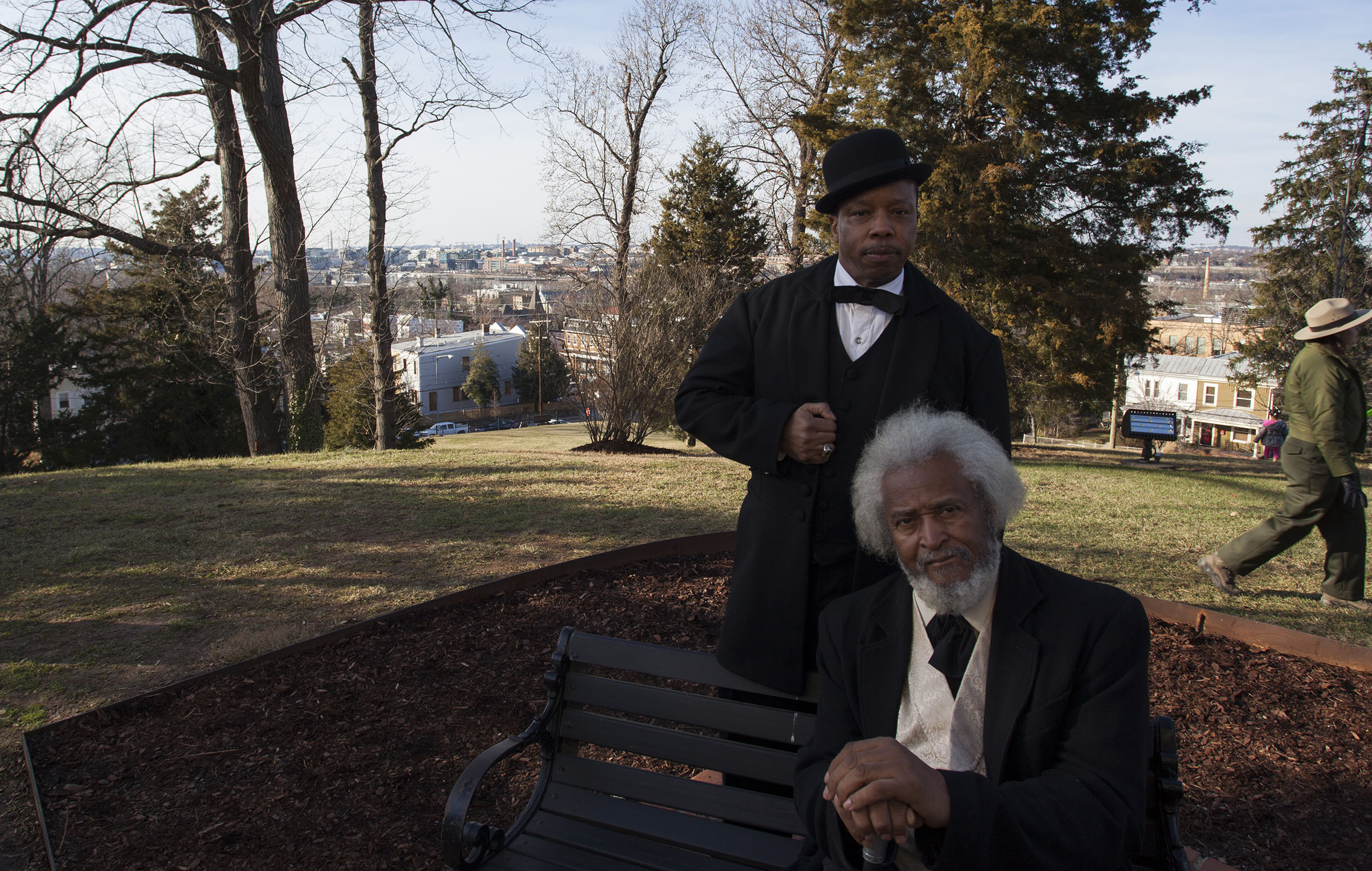 Two African American men in suits, one standing and one seated on a bench, atop a sunny hill with a view of a city beyond.