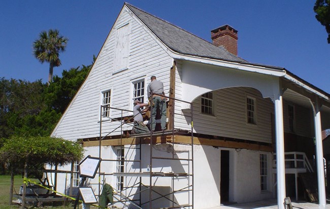 Two NPS staff on scaffolding to work on the second floor exterior of a wooden structure.