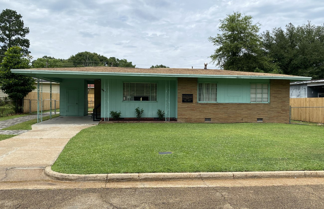 A flat, open lawn connects the road and curb to a one-story ranch house with driveway and carport to the left.