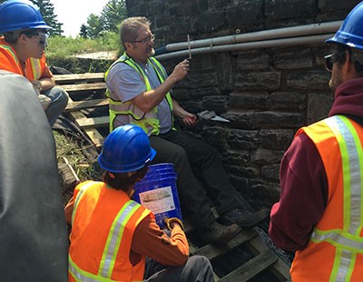 A group of people, wearing safety gear, watch intently as a man holding tools demonstrates maintenance technique beside a stone wall.