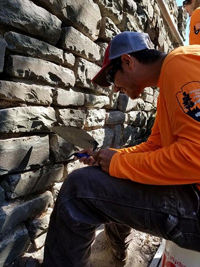 A young man in a baseball cap and sunglasses uses tools to repair mortar in a stone wall.