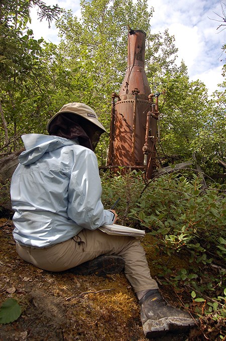 A person seated on the ground amid trees in jacket, hat, and bug net documents a metal mining feature
