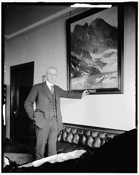 A man in a suit and vest stands in an office next to a leather couch, his arm outstretched to the frame of a painting of a mountain.