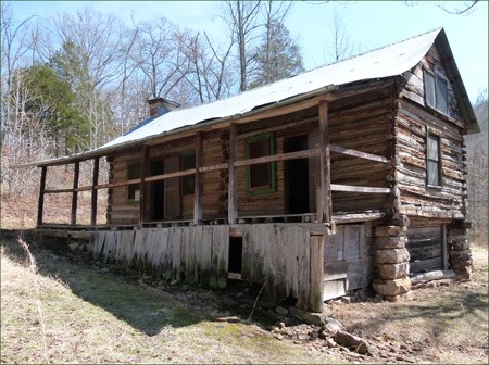 A log cabin with two doors and a long front porch stands on a slope.