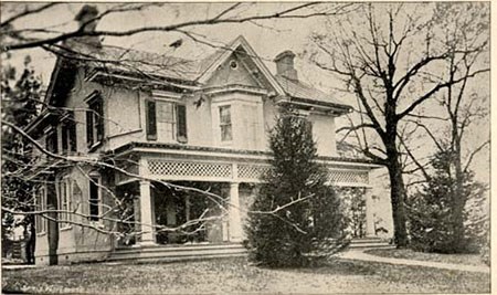 Historic photograph of a two story house, with two young trees at the front and a long front porch.