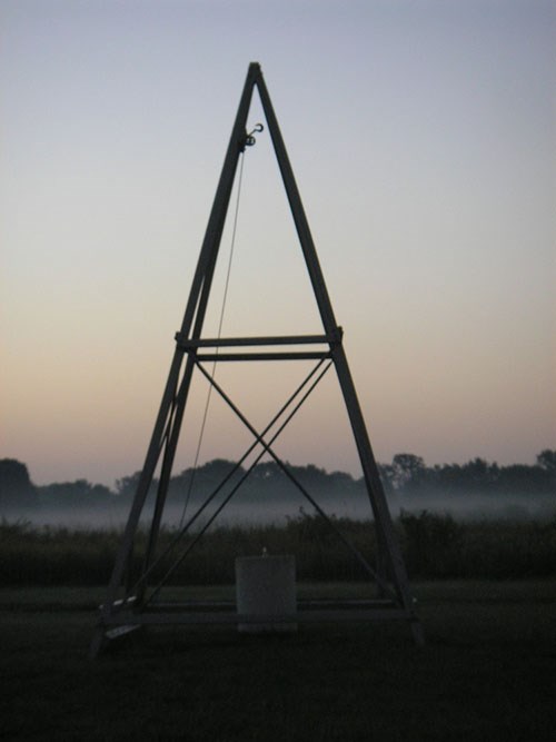Wooden catapult structure shaped like a tall pyramid stands in a hazy field at dawn
