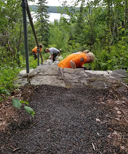 Crew members work on steps on a trail through woods, with a lake in the distance.