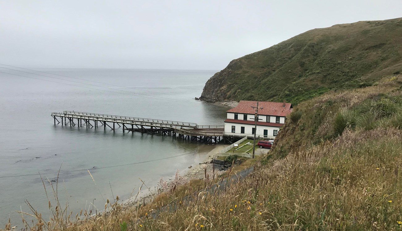 A color photograph of a two-story, white-sided, red-roofed, shore-side building with a wharf/boat ramp that extends to the left into a bay. Steep grassy hills rise beyond and to the right of the boathouse.