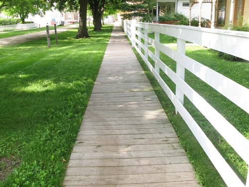 A white fence runs parallel to a wooden walkway, above green grass.