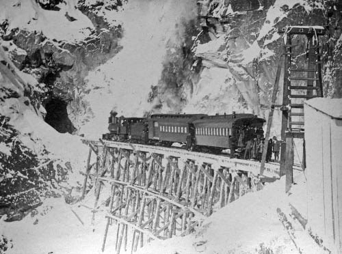 Train on a snowy railroad bridge approaches a tunnel in the side of a mountain.