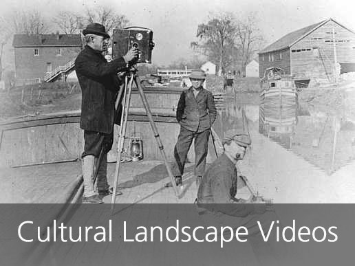 Three men ride a canal boat in the 1920s, one recording on a film camera.