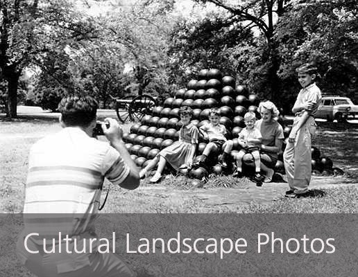 A man kneels to capture a portrait of four kids and a woman, posing on a pile of cannonballs around 1959.