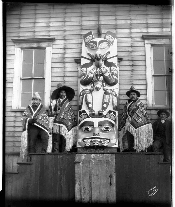 Four men, wrapped in blankets, sit on either side of a totem pole.