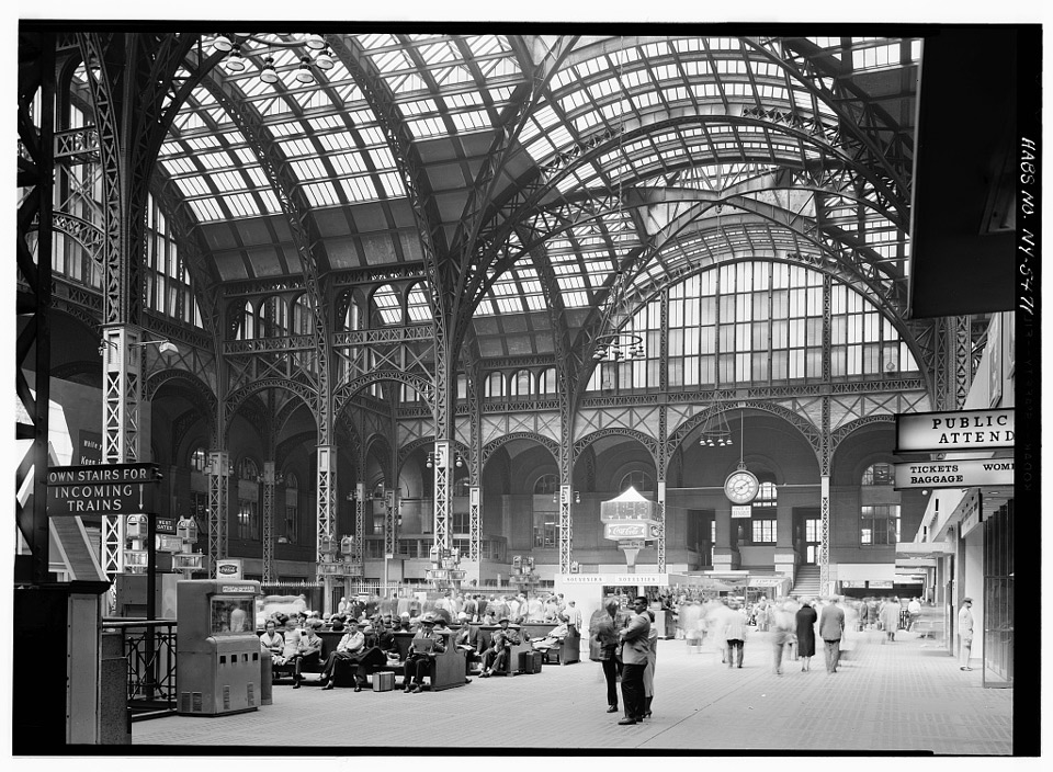 Passengers gather in the expansive concourse of a train station under an arching glass and metal ceiling.