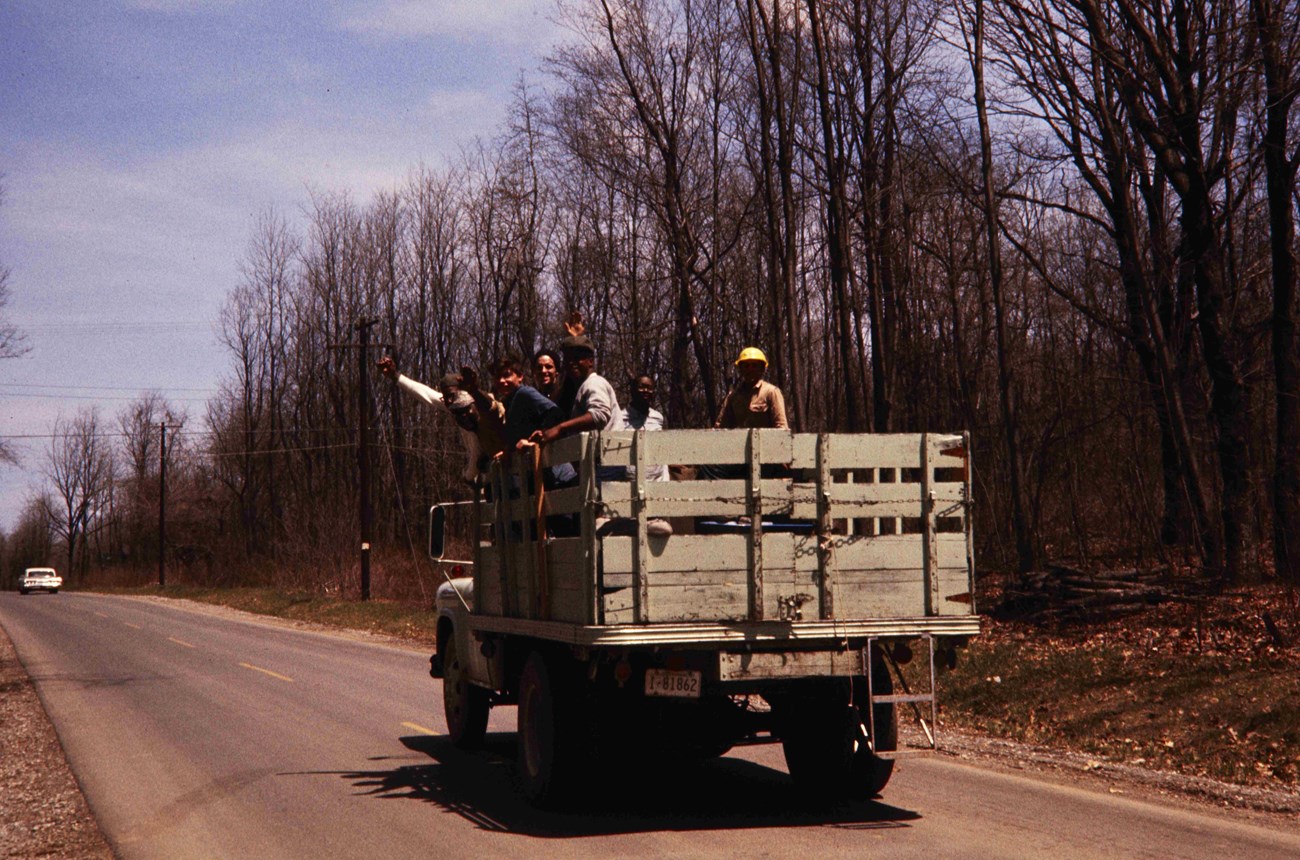 A group of young men wave from a pickup truckbed with wooden sides, on a road.