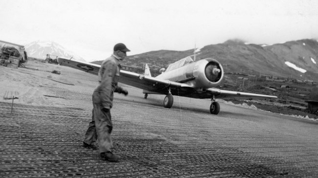 A man in coveralls stands on an airfield in front of a small plane