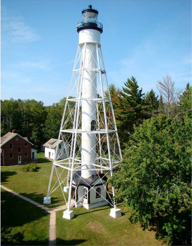 A tall light house with a beacon standing on metal legs, surrounded by a clearing of lawn and pathways