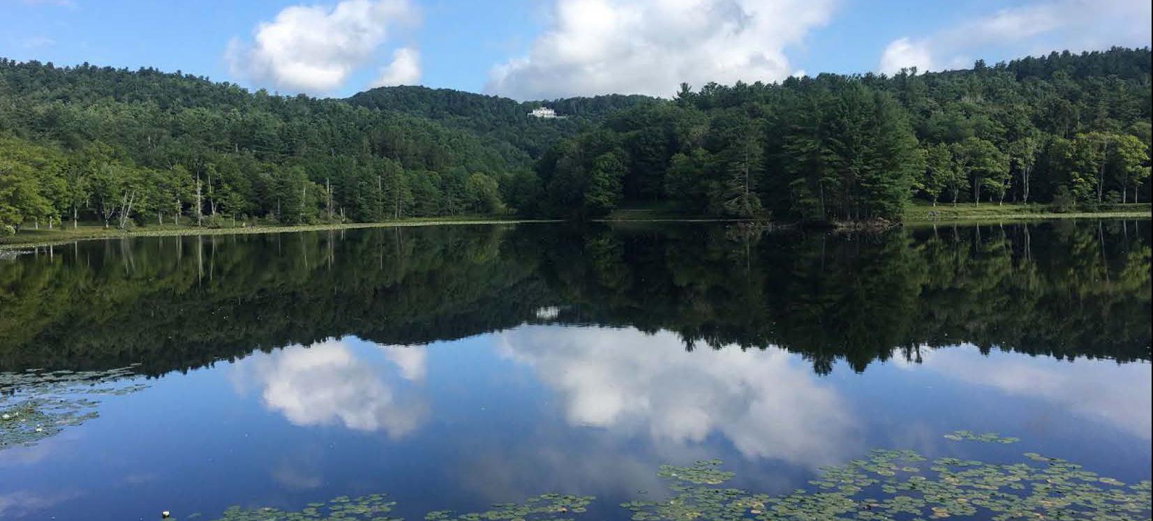 A white house is visible at the top of a distant wooded hillside over a still lake.