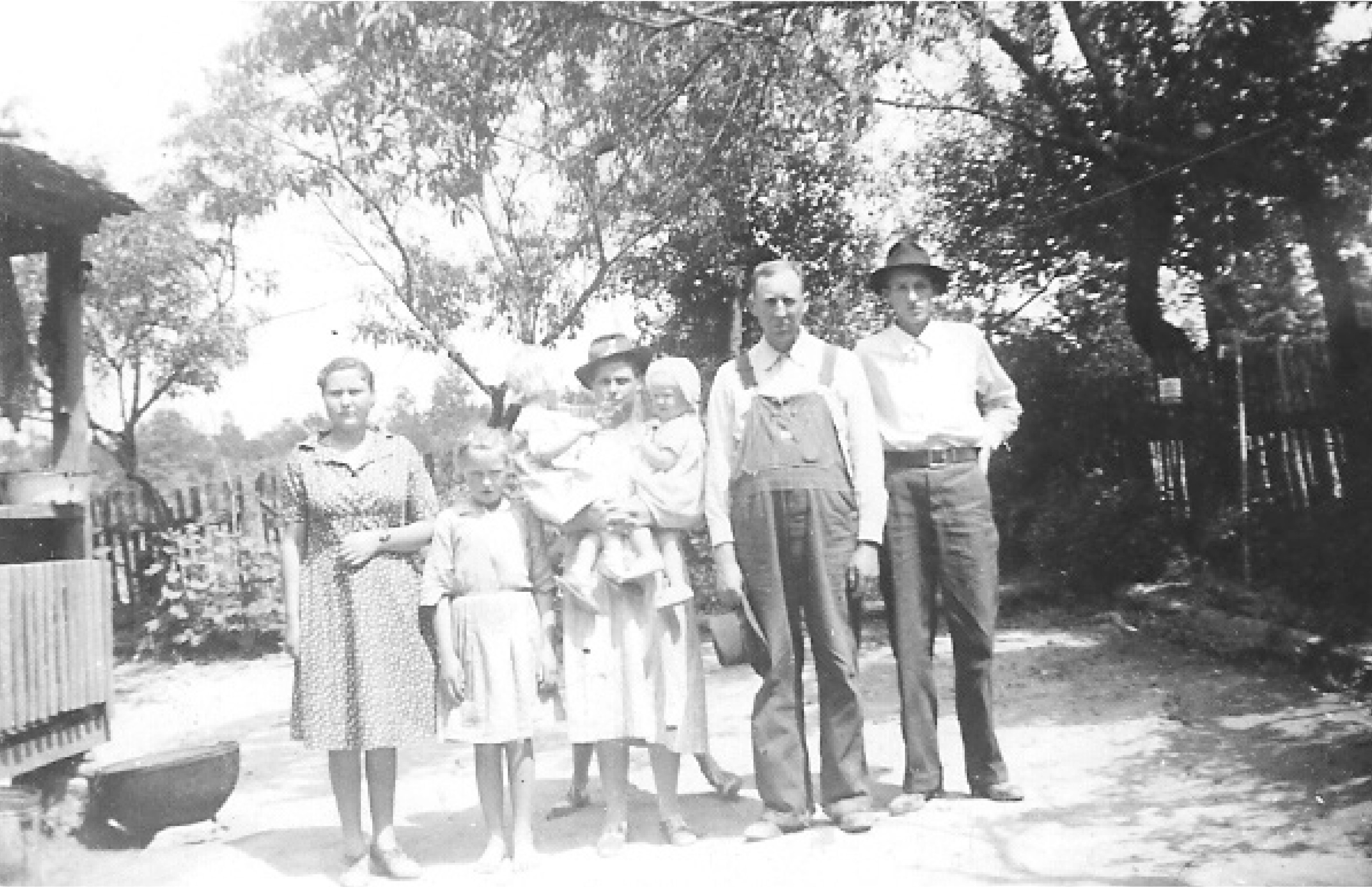 A family of 8 of varying ages in the front yard of a farmhouse, around 1940. Scene includes a swept yard, perimeter yard fence, large trees, and clear vista.