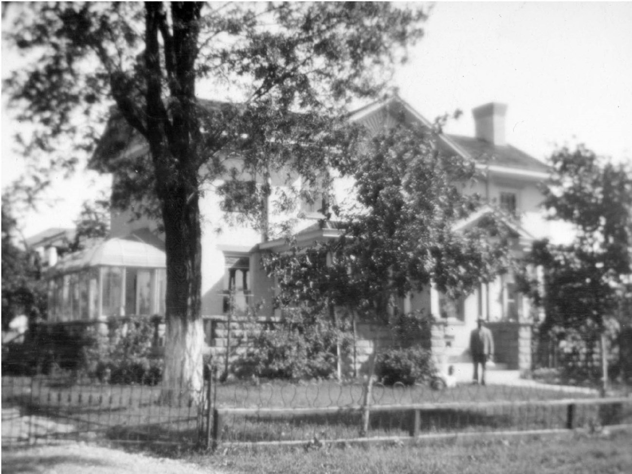 A man stands near the front porch of a two-story house, in a fence-enclosed yard with trees, garden beds,