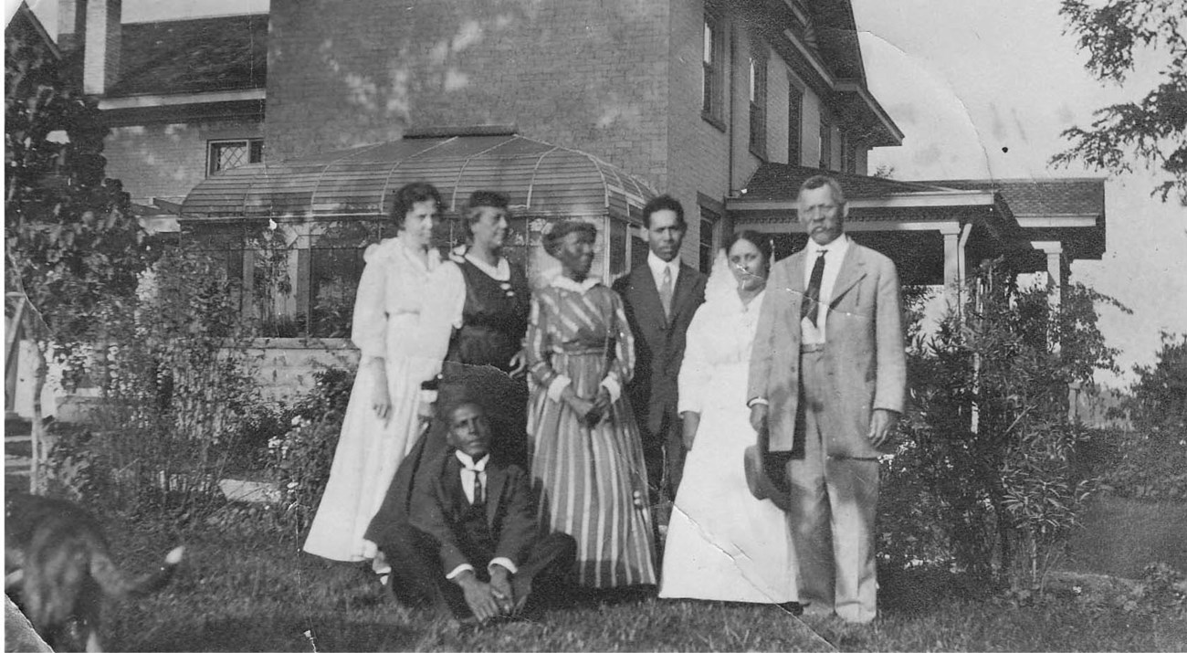 A group of seven people, in dresses and suits, pose beside a row of shrubs. A house with greenhouse is in the background.