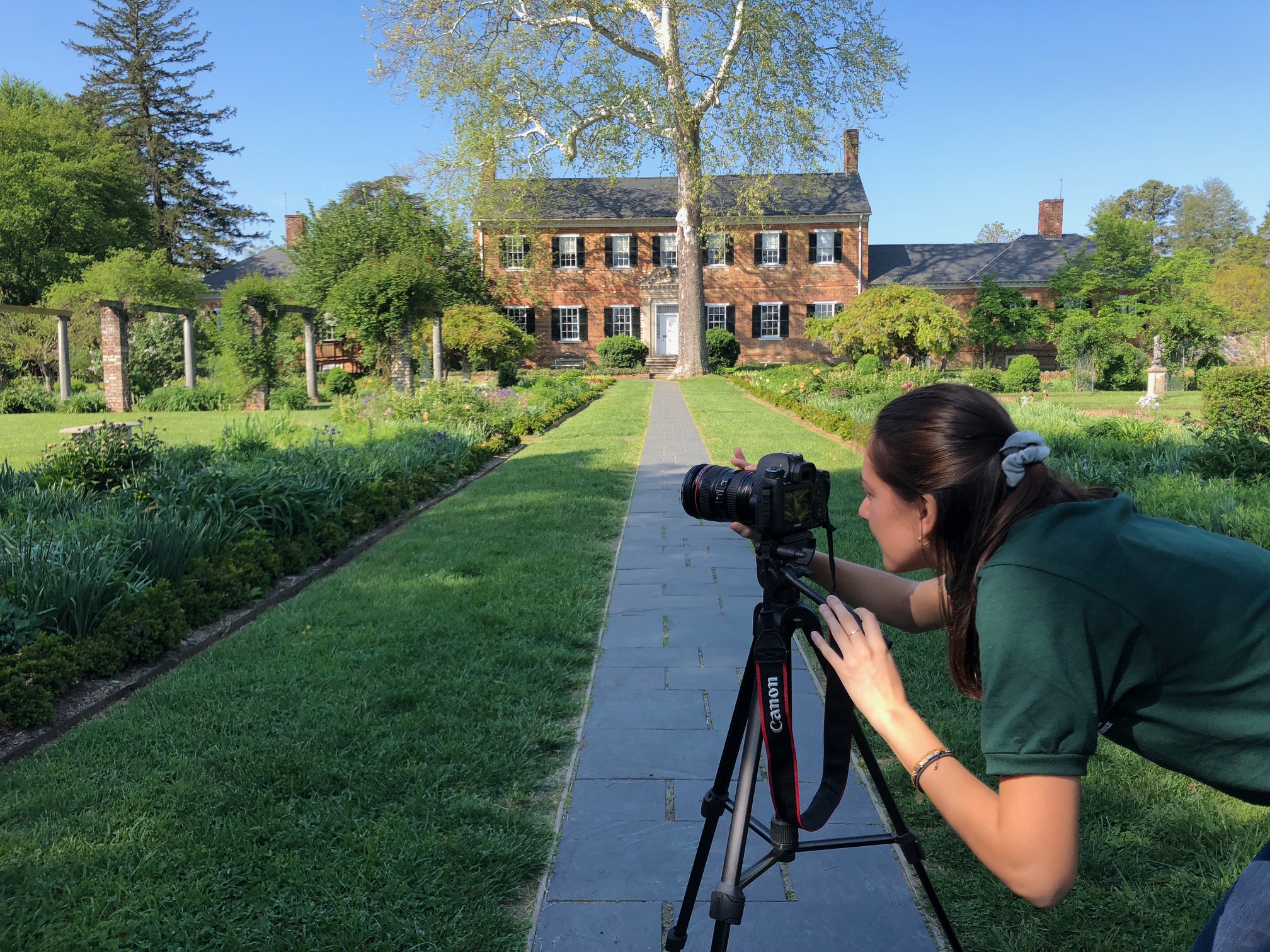 A woman leans over to look through a camera on a tripod, aimed at a formal garden in front of a two story manor house.