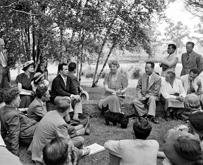 Eleanor Roosevelt sits on a low stone wall, in the center of a gathering of well-dressed UNESCO visitors