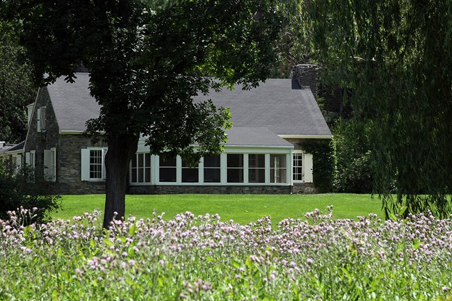 One and a half story stone cottage surrounded by turf, leafy trees, and flowers in the foreground.