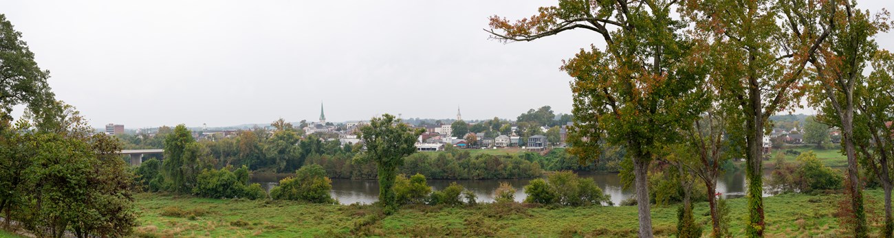 View from a high point across a river bordered by leafy trees to a small town under a cloudy sky.
