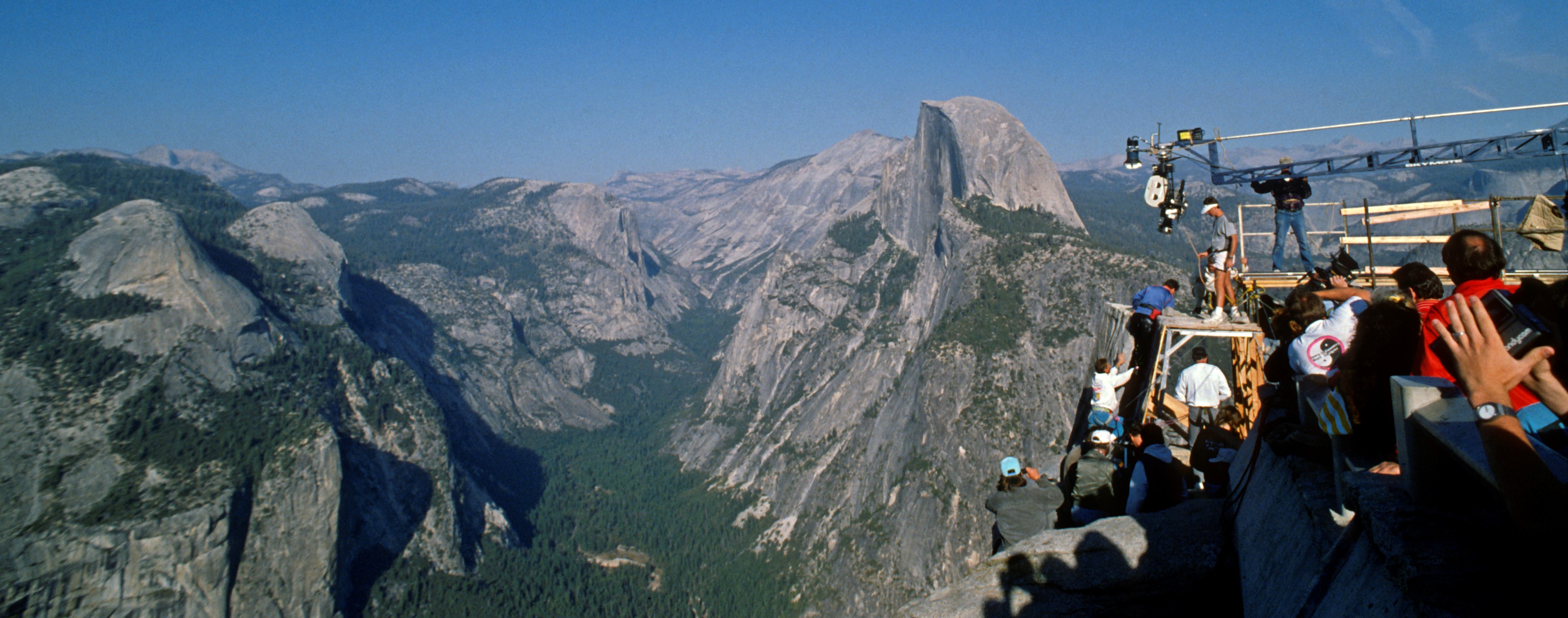 A film crew and cranes is perched at the edge of a deep valley with curving rocks and scattered trees