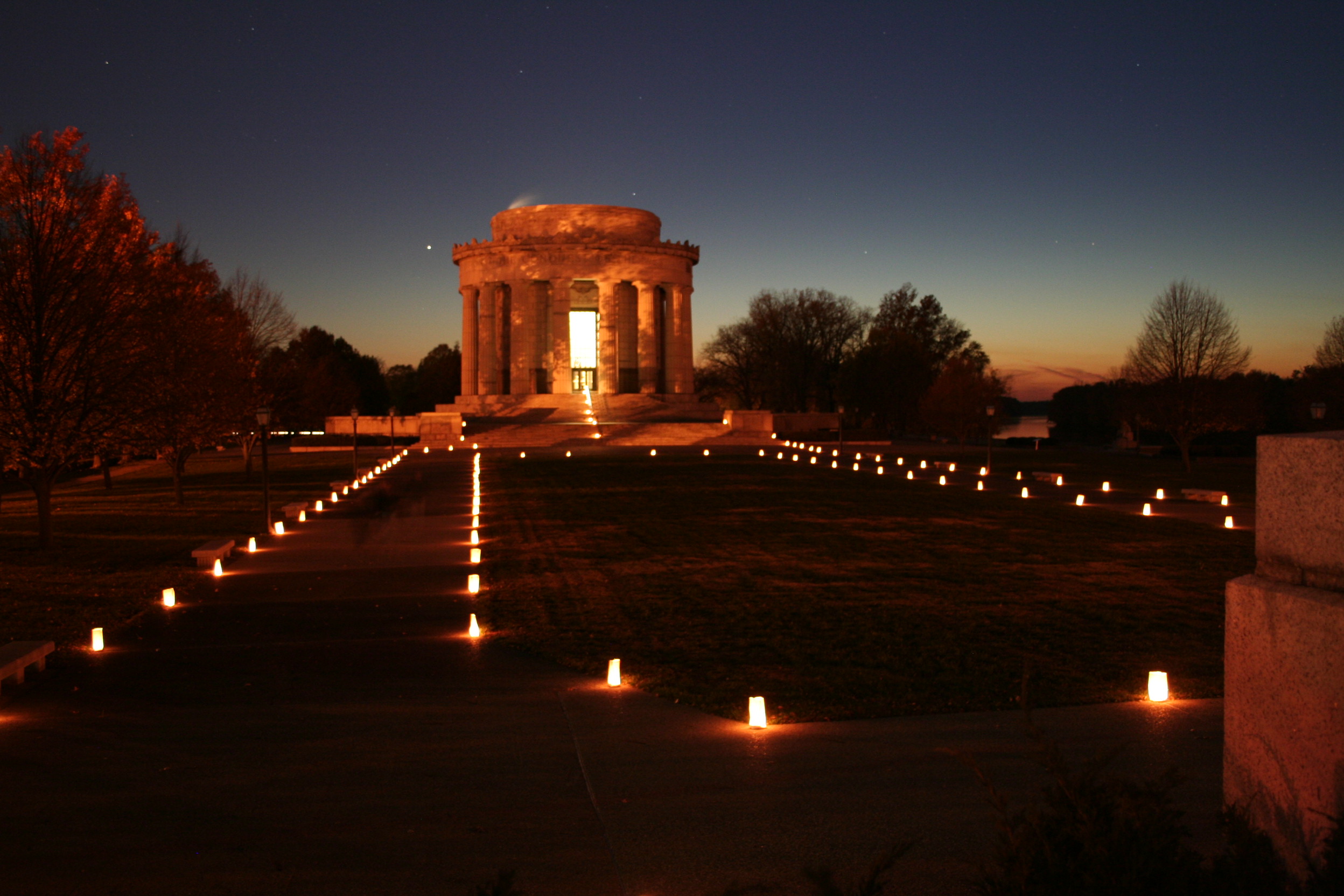 Rows of luminaries lead to a round memorial, surrounded by columns and glowing in the dim twilight
