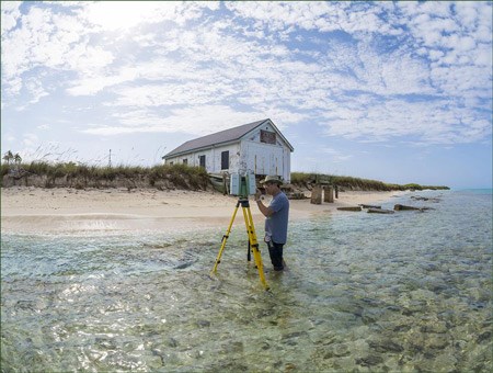A man stands in knee-deep water near a sandy beach, using a laser scanning device on a tripod.