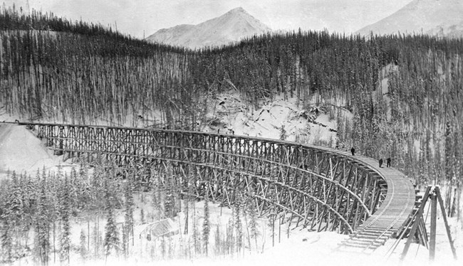 Tiny figures stand on the curve of a long wooden railroad bridge