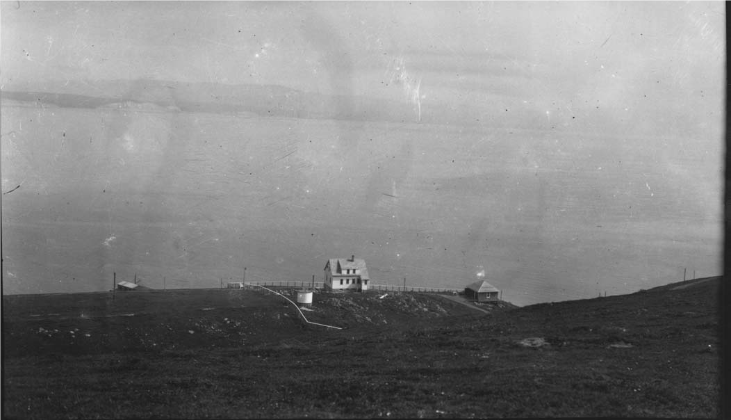 A black and white photograph of a two-story house adjacent to a small two-car garage and a smallish white water tank, all clustered at the base of a grassy slope in the mid-distance above a bay.