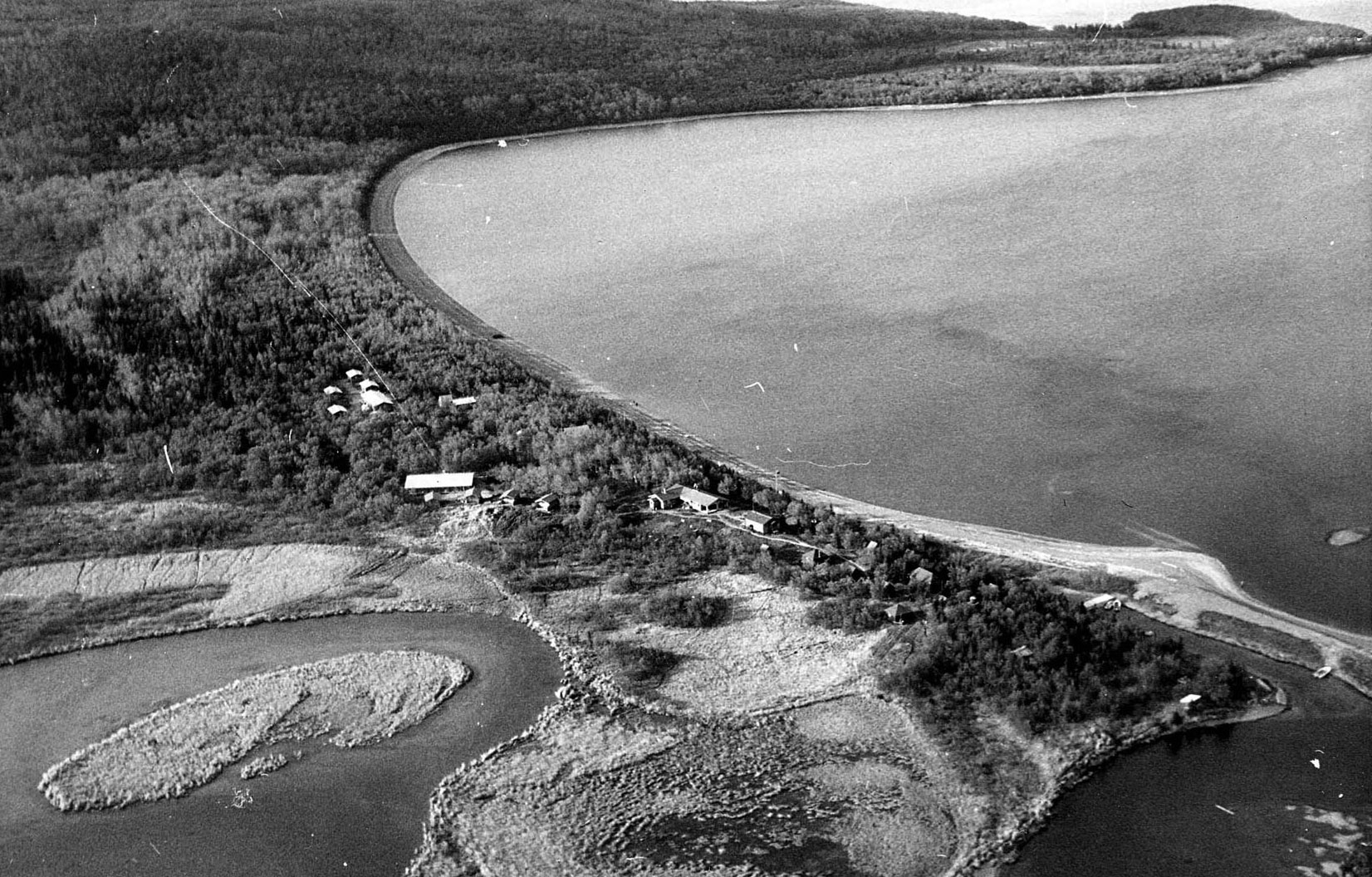 Black and white aerial photo of camp development near a curving shoreline, where a river meets a lake.