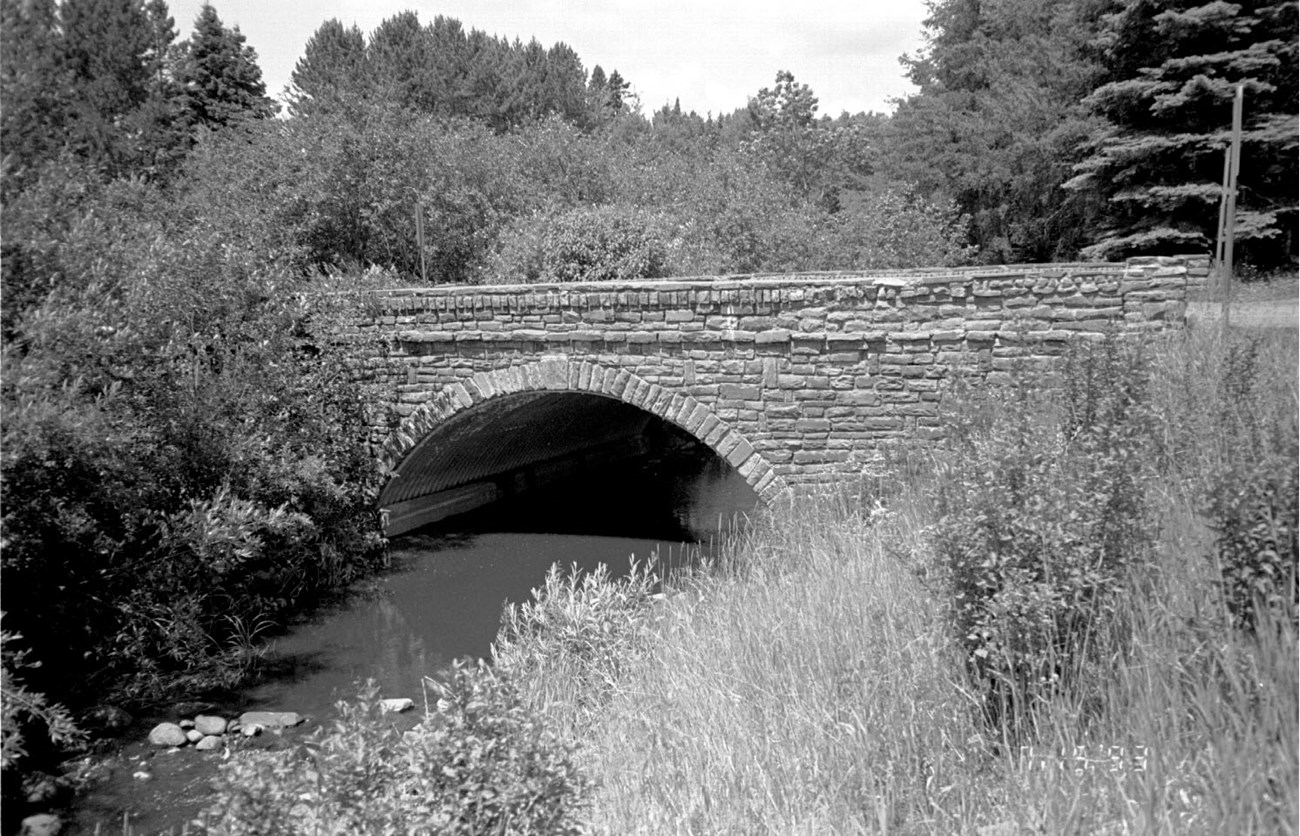 A creek passes under an arching stone bridge, framed by vegetation growing on the banks