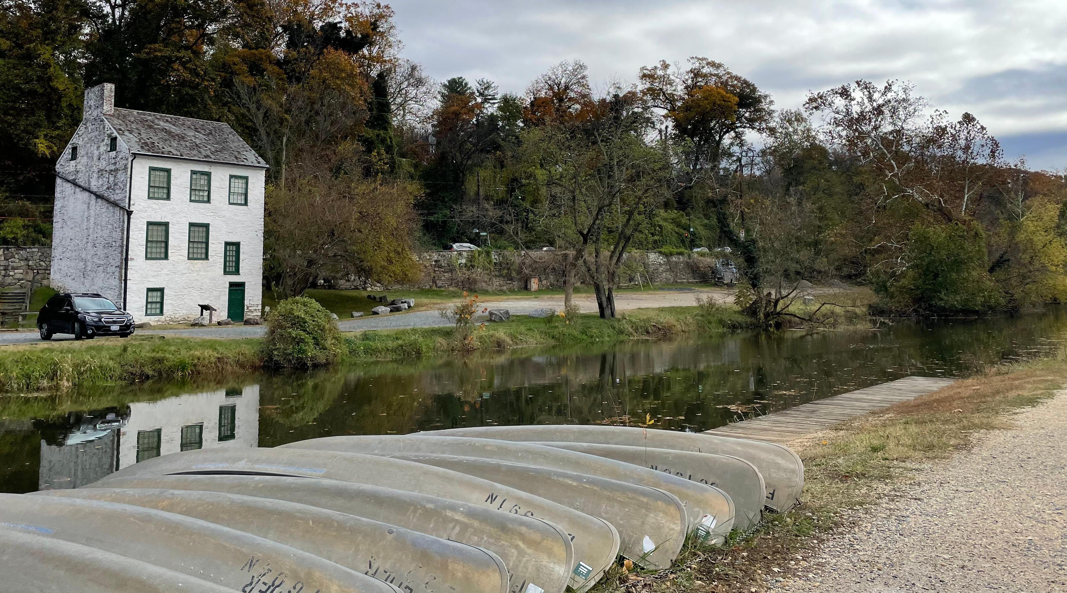 Color photo of the Chesapeake and Ohio Canal and towpath. Several upturned canoes line the shore of the canal in the foreground. The Abner Cloud House and a gravel parking lot can be seen on the opposite shore.
