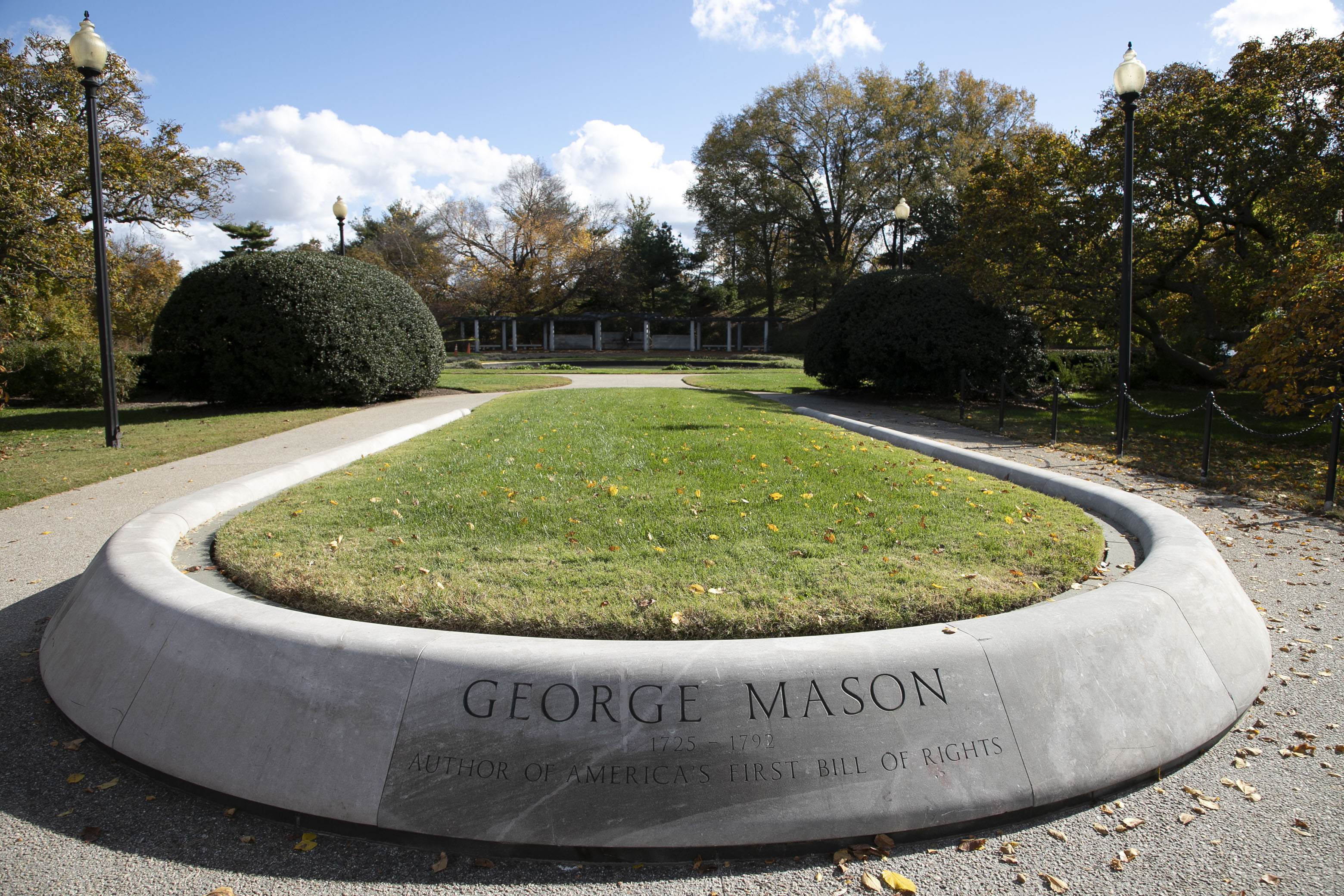 The words “George Mason 1725-1792 Author of America’s First Bill of Rights” are engraved on the edge of a rounded concrete turf planter at the entrance of the George Mason Memorial. A sidewalk, manicured shrubs, and low trees surround the planter.
