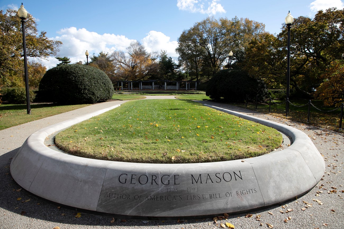 The words “George Mason 1725-1792 Author of America’s First Bill of Rights” are engraved on the edge of a rounded concrete turf planter at the entrance of the George Mason Memorial. A sidewalk, manicured shrubs, and low trees surround the planter.