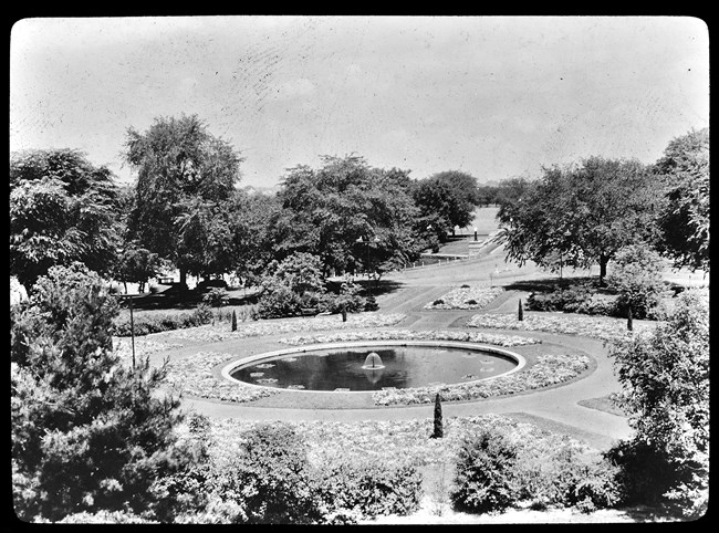 A round fountain is surrounded by a circular walkway and flower beds in the 1930s. A variety of trees line the edge of the garden and Ohio Drive extends into the background.