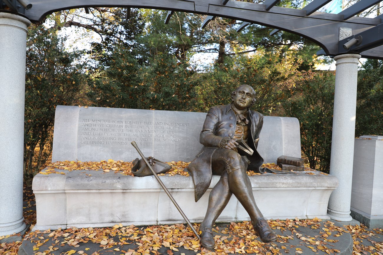 The George Mason statue is a man reclined with crossed legs on a stone bench, holding a book, with tricorn hat, cane, and books on the bench to either side. Stone columns flank the bench and rise to a wooden trellis overhead.