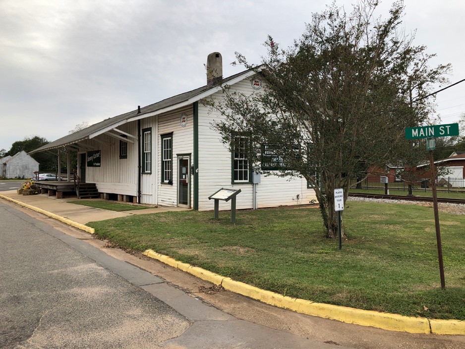 Side angle view of a one-story wooden train depot structure. near a curb and street sign marking "Main St."