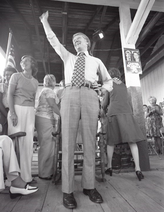 Jimmy Carter smiles and waves, standing among a crowd inside a wooden structure