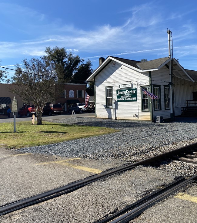 A set of train tracks, gravel, and an area of turf next to a one-story train depot. A sign on the side says "Jimmy Carter Presidential Campaign Headquarters"