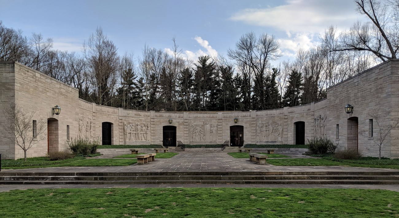 Limestone block building, half circle shape with six openings spaced evenly apart. Light fixtures above four openings. Two windows on each side of left opening and right opening. Green grass in foreground leads to stairs and walkway with stone benches on