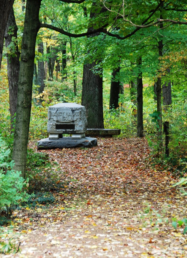 A rectangular stone monument at the end of a trail scattered with leaves, under a canopy of leafy trees