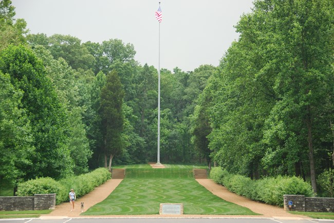 A tall flagpole stands at the end of a neat stretch of turf, lined on each side by rows of leafy trees.